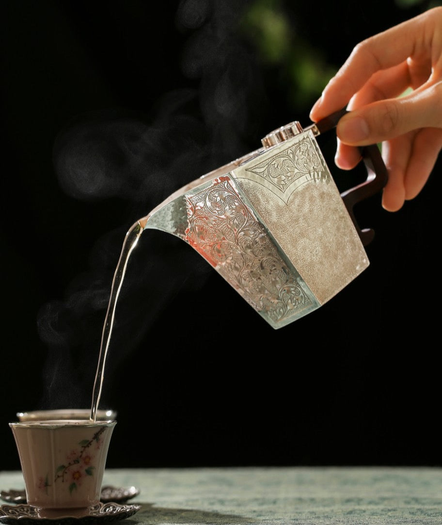 Person pouring steaming hot liquid from a silver teapot into a cup with a blurred green background