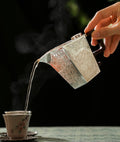 Person pouring steaming hot liquid from a silver teapot into a cup with a blurred green background